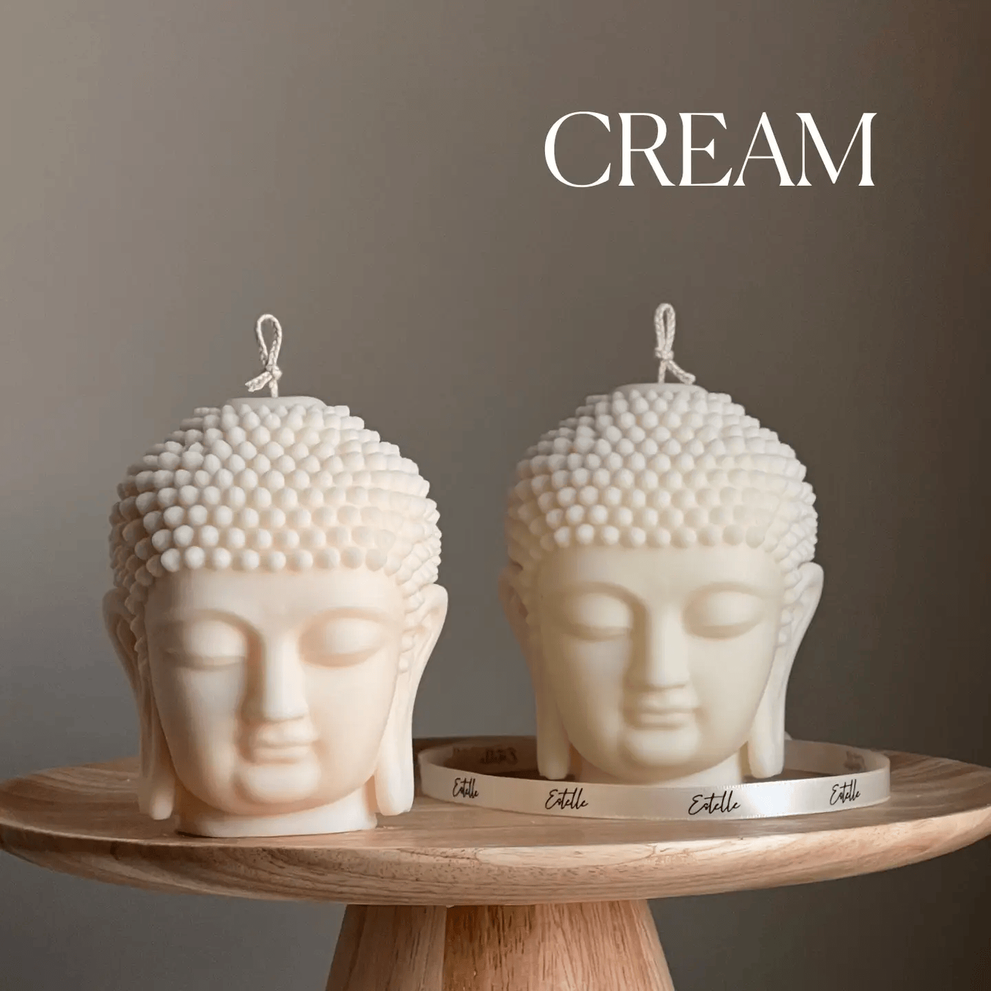 Two cream-colored Buddha head candles on a wooden stand with a neutral background.