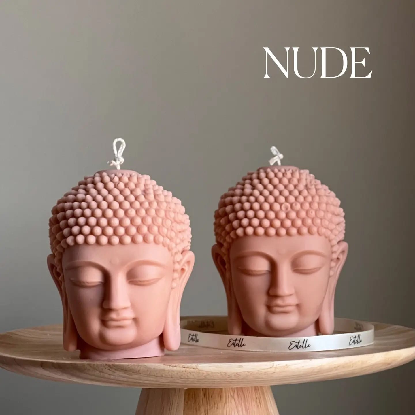 Two pink Buddha head candles on a wooden stand with 'NUDE' text above.