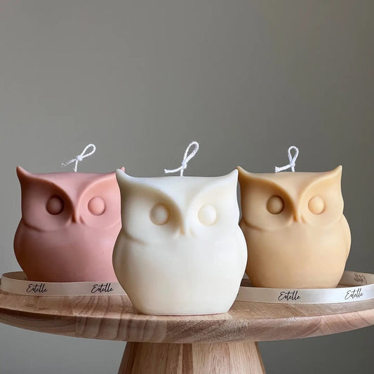 Three owl-shaped candles in pink, white, and beige on a wooden stand with a gray background.