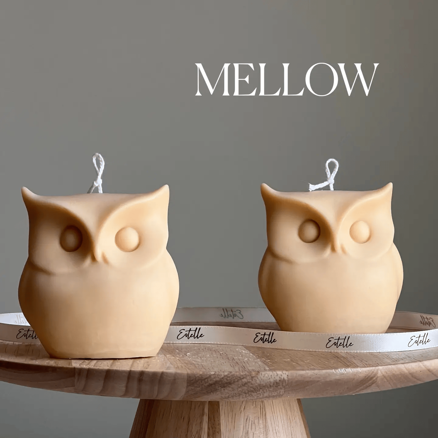 Two owl-shaped candles on a wooden stand with a gray background and 'Mellow' text.