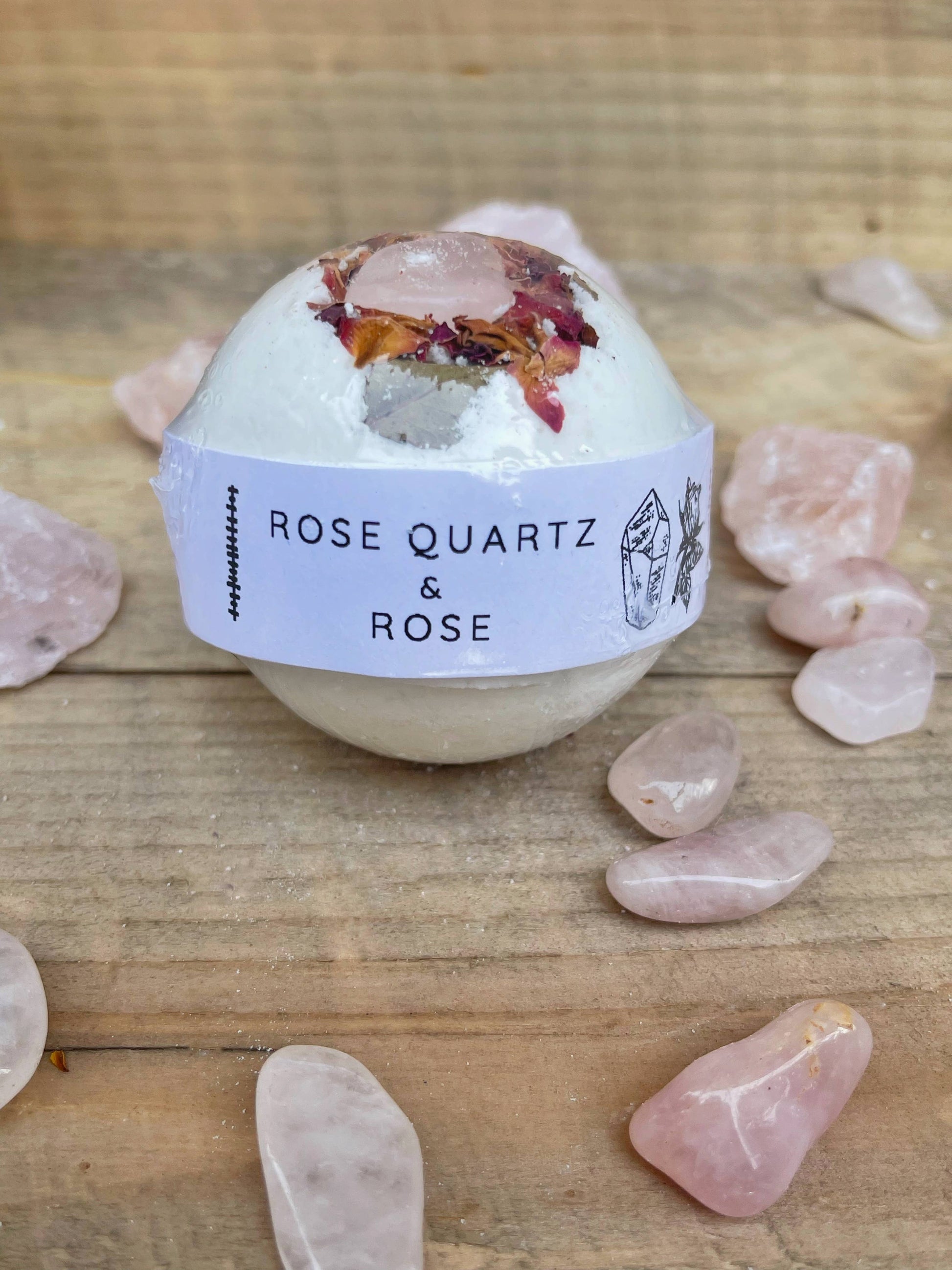 Bath bomb labeled 'Rose Quartz & Rose' surrounded by rose quartz crystals on a wooden surface