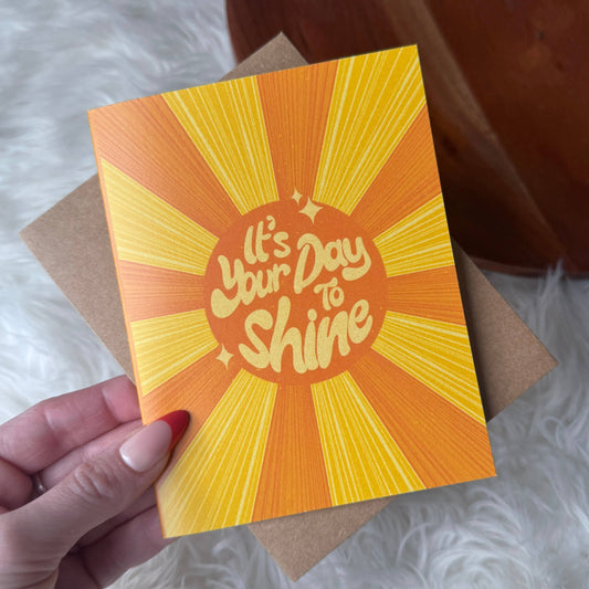 Card with 'It's Your Day to Shine' text on a bright orange and yellow background, held by a hand.