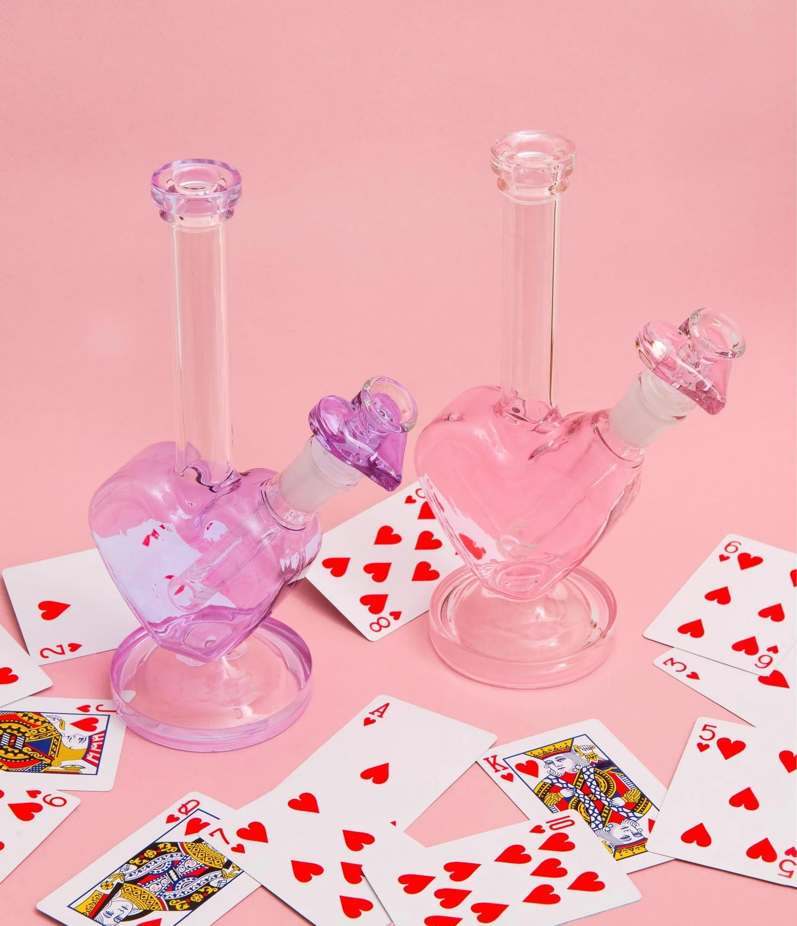 Pink and Purple clear heart-shaped glass smoking devices on a pink background with playing cards.