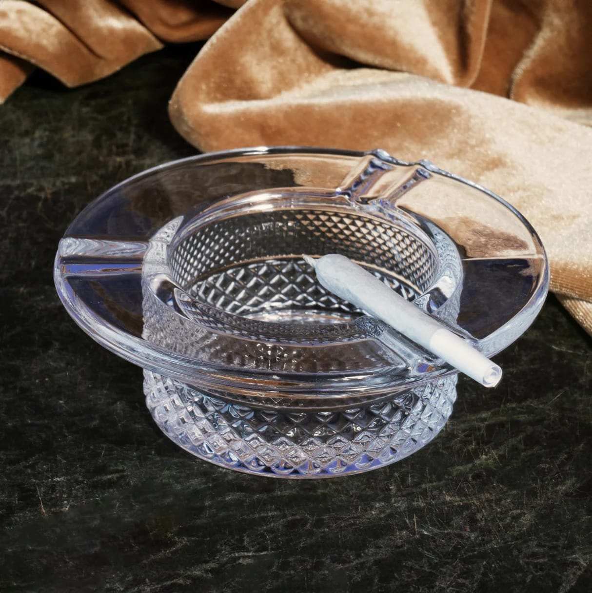 Clear glass ashtray with a textured design on a dark surface with a brown fabric background.