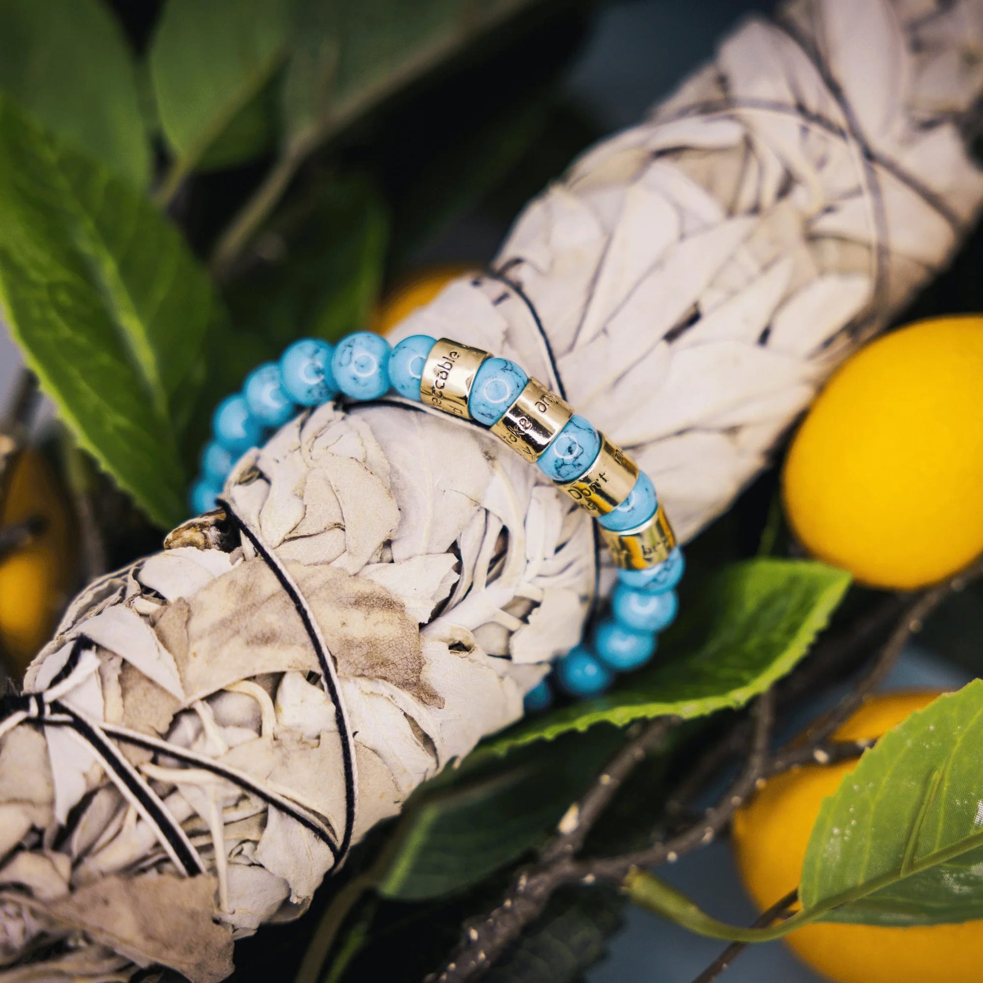Bracelet with blue beads and gold accents on a white smudge stick with green leaves and oranges in the background