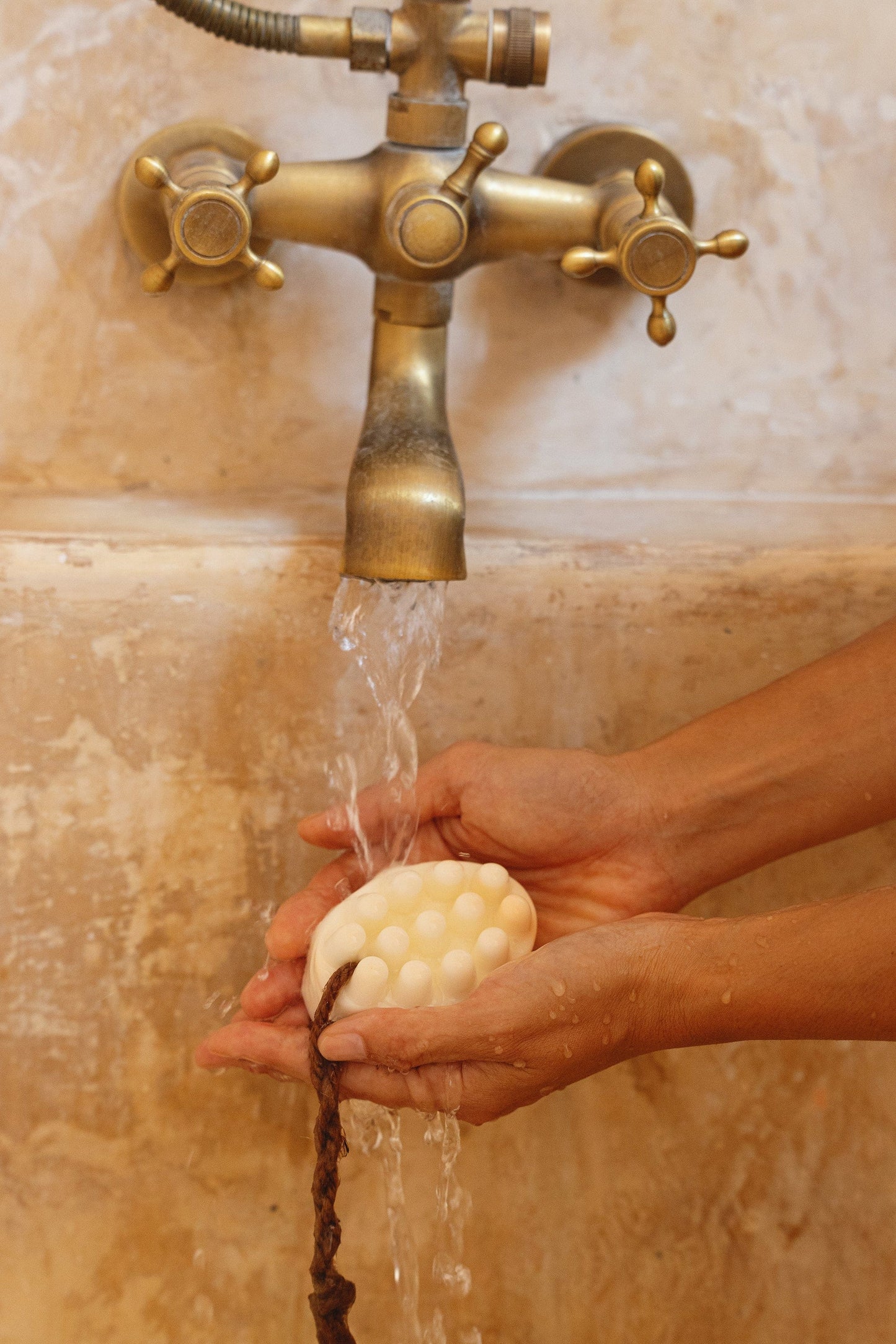 Person washing hands with palo santo soap under a brass faucet on a textured wall.
