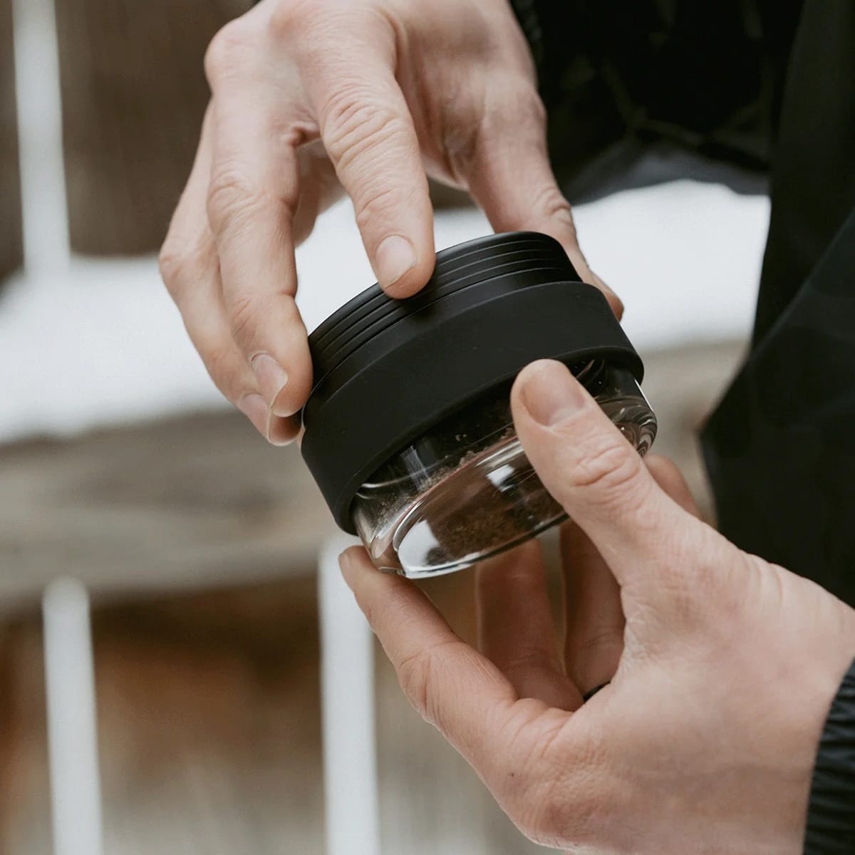 Person holding a glass jar with a black lid, possibly containing coffee grounds.